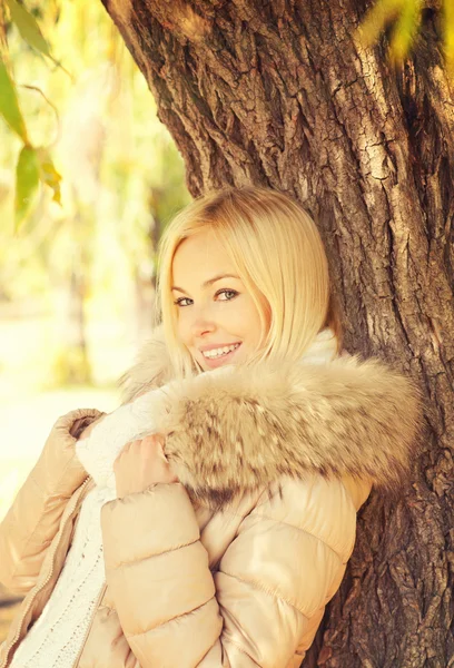 Beautiful elegant smiling blonde woman posing in autumn  park standing near the tree. — Stock Photo, Image