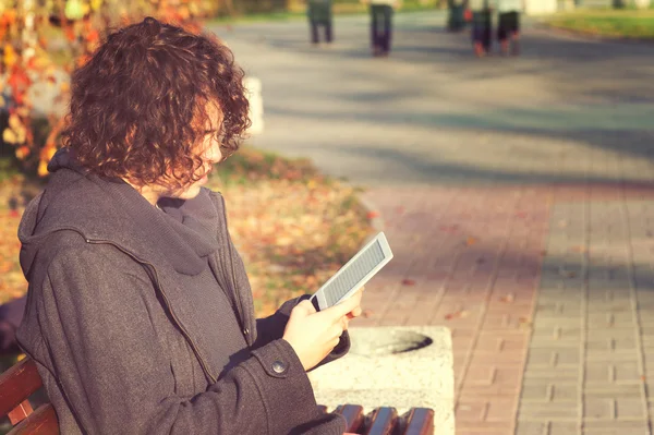 Young man sitting in autumn park and read the electronic book. — Zdjęcie stockowe