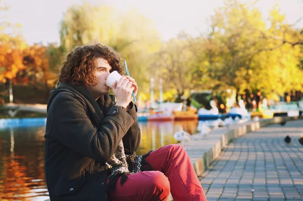 Un tânăr elegant care stă pe malul lacului în parcul de toamnă și bea o cafea . — Fotografie, imagine de stoc