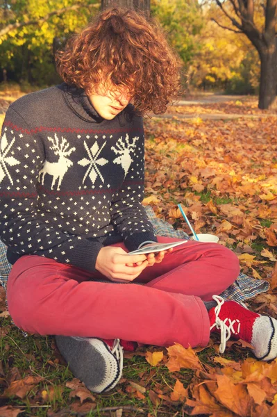 Junger erwachsener Mann sitzt im Herbstpark und liest das elektronische Buch. — Stockfoto