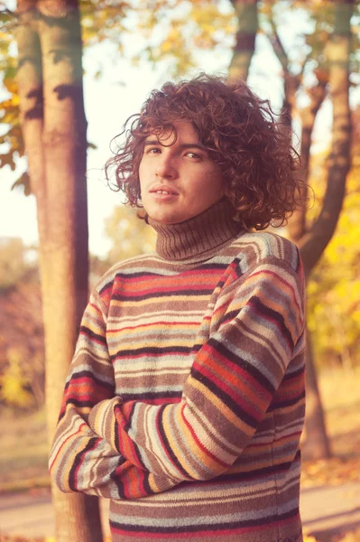 Attractive young man with long curly hair, dressed in striped sweater in autumn park. — Stockfoto