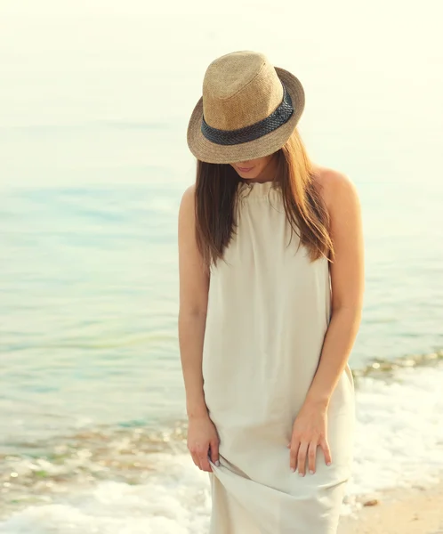 Feliz mujer sonriente caminando en una playa de mar vestida con vestido blanco y sombrero cubriendo la cara, relajándose y disfrutando del aire fresco . — Foto de Stock