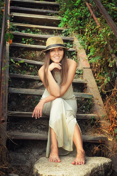 Feliz sorrindo jovem mulher vestida de chapéu e vestido longo branco sentado descalço em uma escada de madeira vintage . — Fotografia de Stock
