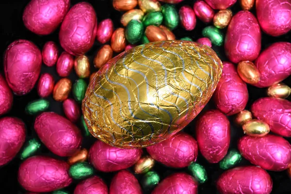 Pile or group of multi colored and different sizes of colourful foil wrapped chocolate easter eggs in pink, red, gold and lime green with a large gold milk chocolate egg in the middle.