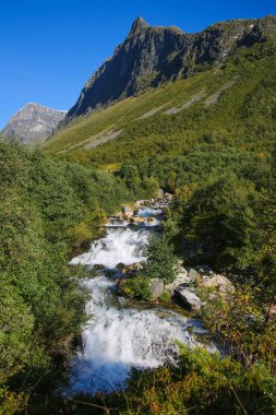 Beautiful views from Fossersa hiking trail of Storsterfossen waterfall with mountains, and valley landscape in the background. Close to Geiranger fjord, Norway. Summers day with clear blue sky over the scenery. clipart