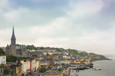 İrlanda, Cobh limanı ve kasaba manzarası. Yamaçta renkli binalar var. Şehrin yukarısında denize bakan büyük bir kilise var. Rıhtım boyunca dolaş. Mantar Portu. 