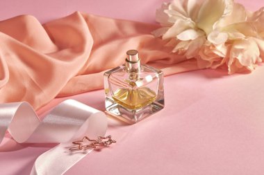 Pembe arka planda bir parfüm şişesi, parçalanmış kumaş, beyaz saten kurdele ve yıldız şeklinde saç tokası. Bahar teması