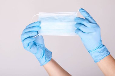 Gri arka planda solunum cerrahisi maskesi tutan tek kullanımlık mavi eldivenli eller. Pandemik korona virüsü, sigorta, hava hastalıkları, SARS, grippe. 