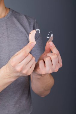 İşitme cihazı tutan adam, yakın plan. Gri arka planda işitme cihazları, ameliyata alternatif. Seçim konsepti