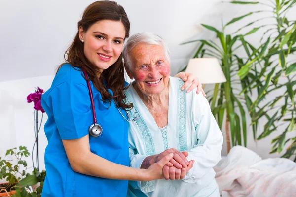 Happy senior kvinna om i sängen med en omtänksam sjuksköterska utövar tillsyn över — Stockfoto