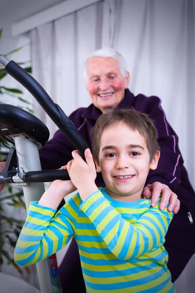 Abuela entrenando en casa en una bicicleta estacionaria con su nieto —  Fotos de Stock