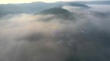 Güneşin doğuşundaki köyün sisli havadan görünüşü. Sabahları orman ve kilisenin üzerinde uçmanın muhteşem manzarası. Sihirli sis. Hava atışı, 4K