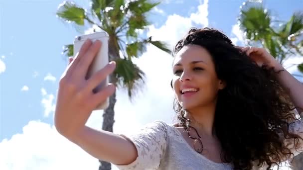Retrato de uma jovem bonita tirando selfie com um telefone inteligente contra o céu azul e as palmas das mãos, câmera lenta — Vídeo de Stock