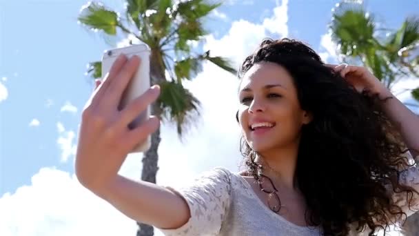 Retrato de una hermosa joven tomando selfie con un teléfono inteligente contra el cielo azul y las palmas, cámara lenta — Vídeos de Stock