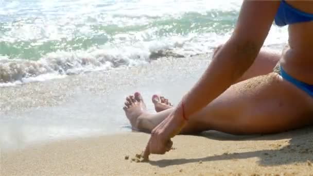 Γυναίκα χαλαρώνοντας στην παραλία, σχέδιο καρδιά στην άμμο, κύματα εκτοξεύεται στο σώμα της, τα πόδια μόνο — Αρχείο Βίντεο