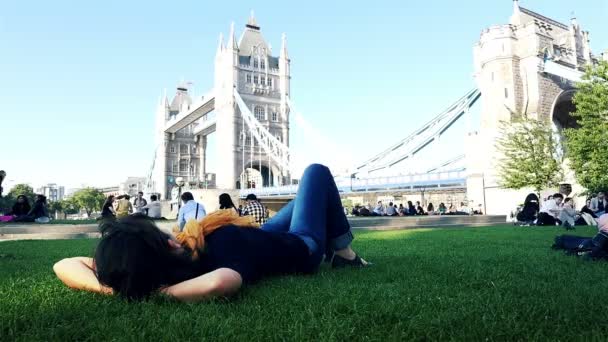 Mulher está deitado em um gramado por Tower Bridge, lapso de tempo de pessoas se movendo ao longo do caminho do Tamisa — Vídeo de Stock