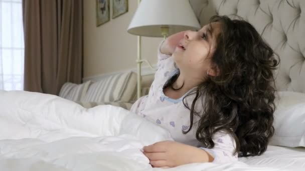 Симпатичная девушка в ночной рубашке, болтающая по мобильному телефону, лежащему на кровати — стоковое видео