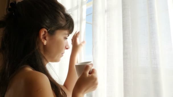 Donna in camicia da notte sorseggiando il suo caffè mattutino dalla finestra e guardando fuori — Video Stock