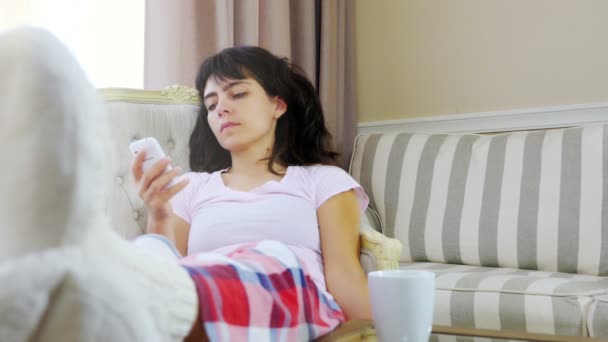 Mujer en pijama está hablando por teléfono móvil en casa, una taza de café en la mesa — Vídeos de Stock