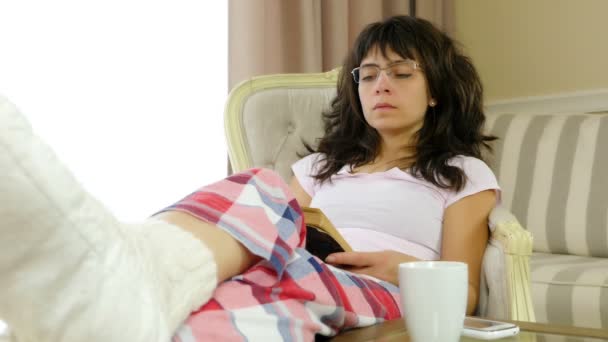 Kobieta w piżamie czyta książkę w domu, filiżankę kawy i telefon komórkowy na stole. — Wideo stockowe