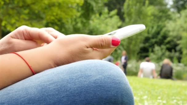 Крупним планом жінка, використовуючи мобільний телефон, сидячи в парку, тільки руки — стокове відео