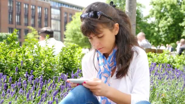 Vrouw die een smartphone gebruikt terwijl ze in een park in Londen zit — Stockvideo