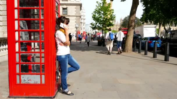 Kvinna med smart telefon samtidigt lutande på en röd telefonkiosk i London, människor gå omkring henne — Stockvideo