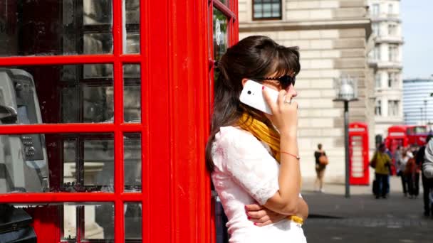 Kvinna med smart telefon samtidigt lutande på en röd telefonkiosk i London, människor gå omkring henne — Stockvideo