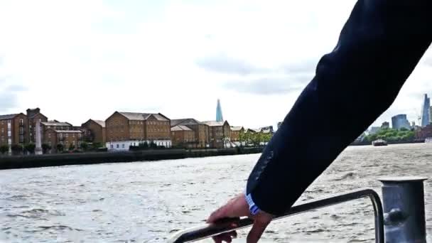 Egy üzletember ruháját fújja a szél egy hajón a Temze folyón, London, Tower híd hátul. — Stock videók