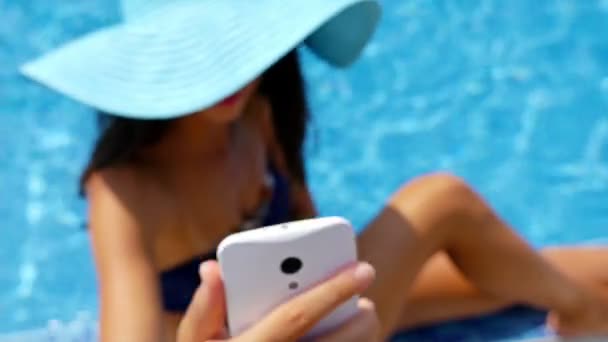 Mujer joven en traje de baño y sombrero azul sentada en el borde de una piscina y tomando una foto de sí misma con teléfono celular — Vídeos de Stock