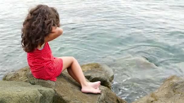 Little girl in red summer dress standing on the rocks on the sea shore and looking to the sea — Stock Video