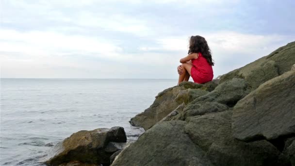 Kis lány piros nyári ruha, a sziklák, a tenger partján állt, és a tengerre néz — Stock videók