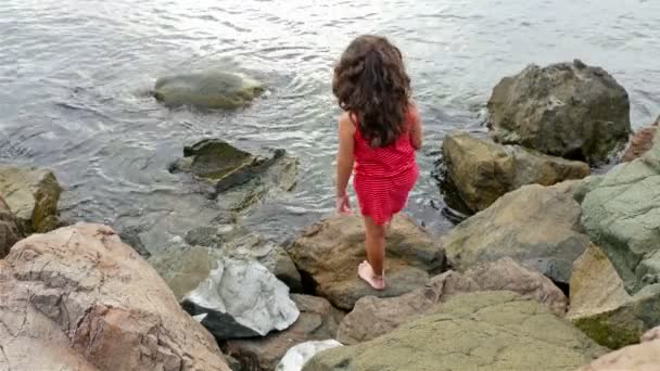 Niña en vestido rojo de verano de pie sobre las rocas en la orilla del mar y mirando al mar — Vídeos de Stock