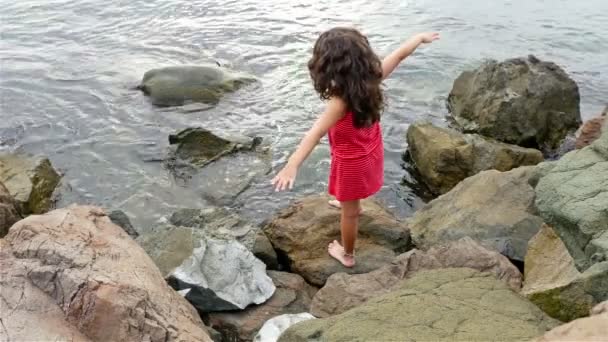Маленька дівчинка в червоній літній сукні стоїть на скелях на березі моря і дивиться на море — стокове відео