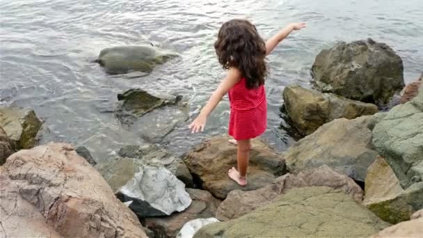 Lilla flickan i röd sommarklänning stående på klipporna vid havet och tittar till havet — Stockvideo