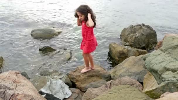 Niña en vestido rojo de verano de pie sobre las rocas en la orilla del mar y mirando al mar — Vídeos de Stock