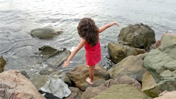 Niña en vestido rojo de verano de pie sobre las rocas en la orilla del mar y mirando al mar — Vídeos de Stock