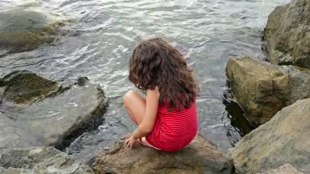 Kleines Mädchen in rotem Sommerkleid, das auf den Felsen am Ufer des Meeres steht und zum Meer blickt — Stockvideo