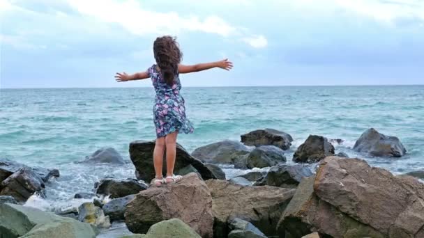 Liten flicka i en klänning på klipporna vid havet och ser att det stormande havet — Stockvideo