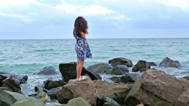 Klein meisje in een jurk staande op de rotsen aan de zee oever en kijken naar de stormachtige zee — Stockvideo