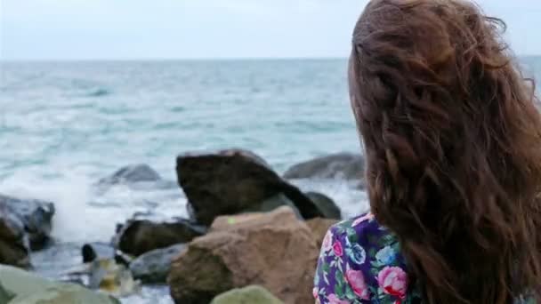 Kleines Mädchen in einem Kleid, das auf den Felsen am Ufer des Meeres steht und auf die stürmische See blickt — Stockvideo