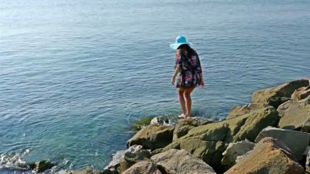 Mujer joven en vestido de verano y sombrero azul sentado en las rocas en la orilla del mar — Vídeos de Stock