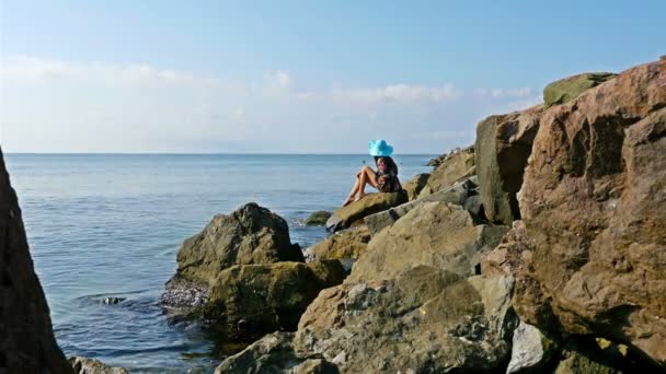 Young woman in summer dress and blue hat sitting on rocks on the sea shore — Stock Video