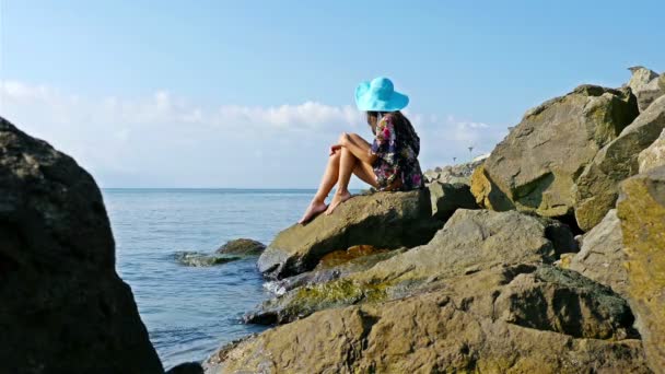 Jonge vrouw in zomerjurk en blauwe hoed zittend op de rotsen aan de zee oever — Stockvideo
