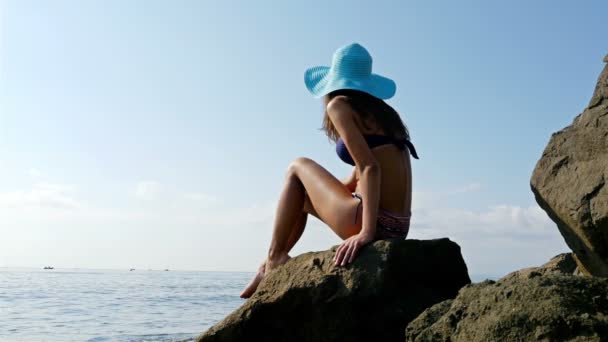 Jonge vrouw in siwmming pak en blauwe hoed zittend op de rotsen aan de zee oever — Stockvideo