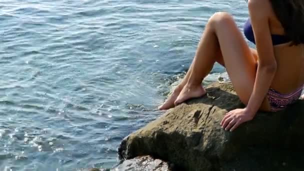 Молодая женщина в купальнике и синей шляпе сидит на скалах на берегу моря — стоковое видео