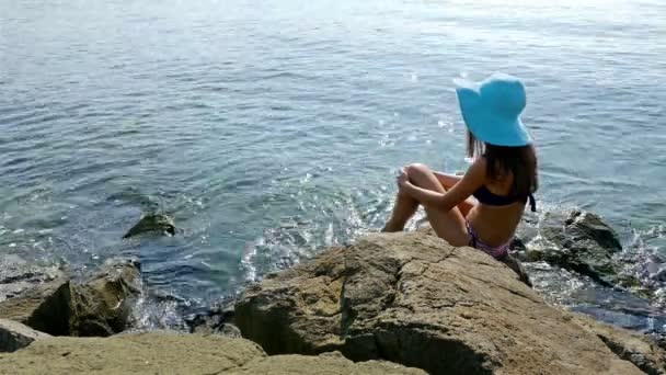 Jeune femme en maillot de bain et chapeau bleu assis sur des rochers au bord de la mer — Video
