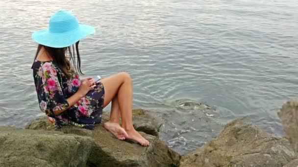 Mujer joven en un vestido de verano y sombrero azul sentado en la costa rocosa del mar y tomar una foto de sí misma con el teléfono celular — Vídeos de Stock