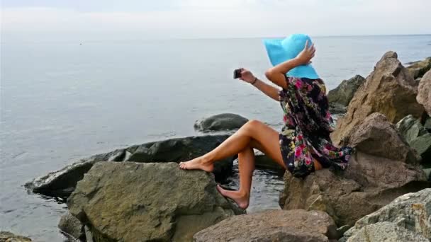 Young woman in a summer dress and blue hat sitting on the rocky sea shore and taking photo of herself with cellphone — Stock Video