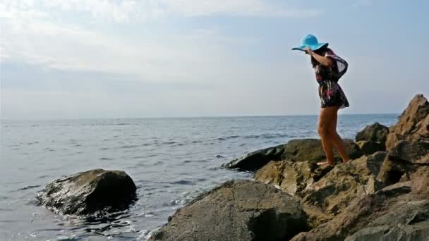 Junge Frau im bunten Sommerkleid mit Blick auf das Meer am felsigen Ufer — Stockvideo